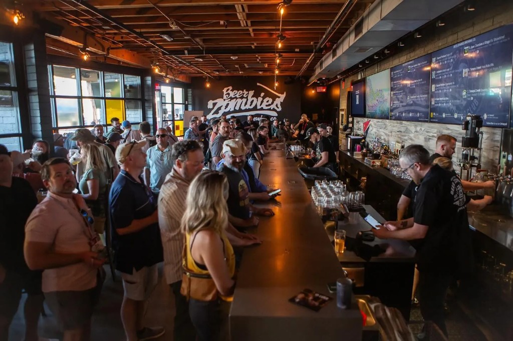 A packed taproom bar with the tap list on screens overhead
